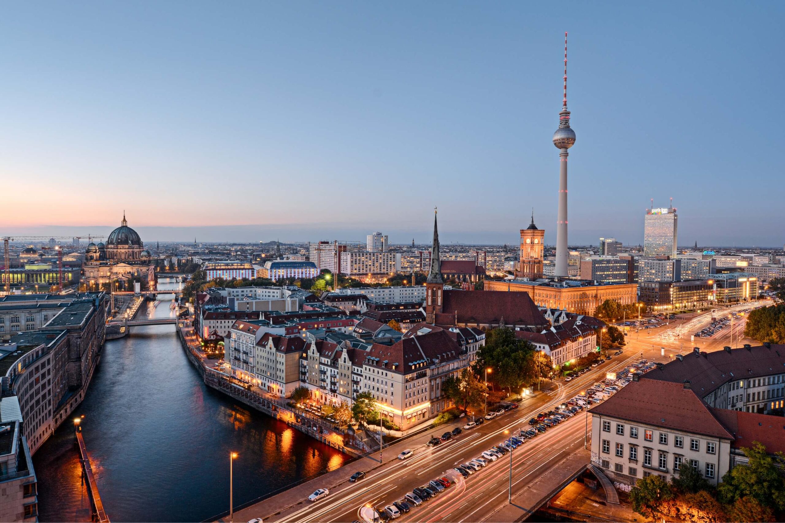 Weitwinkelaufnahme der Berliner Skyline bei Dämmerung. Im Zentrum des Bildes ragen der Berliner Fernsehturm und der Berliner Dom empor. Die Spree schlängelt sich durch die Stadt und reflektiert die Lichter der Gebäude. Mehrere Brücken überspannen den Fluss. Die Straßen sind von Autos befahren. Der Himmel ist in verschiedenen Blautönen gefärbt, während die Lichter der Stadt bereits eingeschaltet sind.