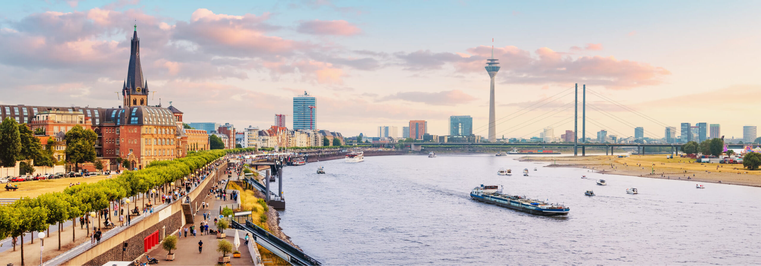 Weitwinkelaufnahme der Düsseldorfer Skyline bei Tageslicht. Der Rhein fließt im Vordergrund und teilt die Stadt. Mehrere Schiffe und Boote sind auf dem Wasser unterwegs. Die Oberkasseler Brücke überspannt den Fluss. Im Hintergrund erhebt sich der Rheinturm. Die Altstadt mit ihren historischen Gebäuden und dem markanten Kirchturm ist auf der linken Seite des Bildes zu sehen. Die Rheinpromenade ist von Menschen belebt. Der Himmel ist mit einigen Wolken bedeckt.