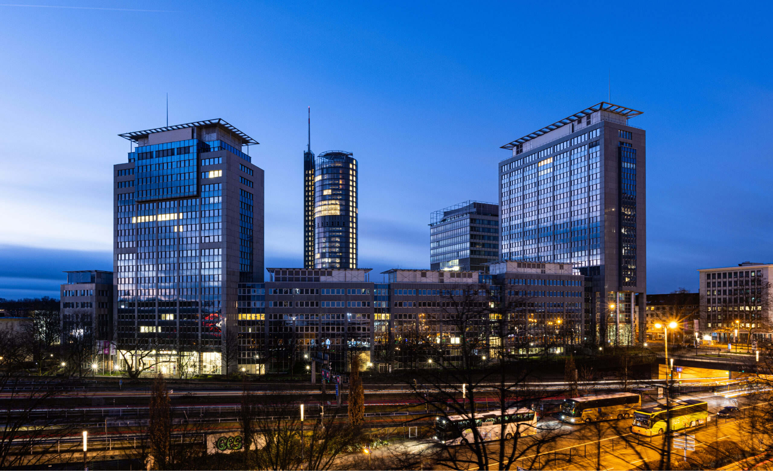 Panoramaaufnahme der Essener Skyline bei Dämmerung. Im Vordergrund sind Bahngleise mit vorbeifahrenden Zügen und Bussen zu sehen. Im Hintergrund erheben sich mehrere beleuchtete Hochhäuser mit blauer Glasfassade und flachen Dächern. Die Straßenbeleuchtung und die Lichter in den Fenstern der Gebäude erhellen die Szene. Der Himmel ist blau mit vereinzelten Wolken.