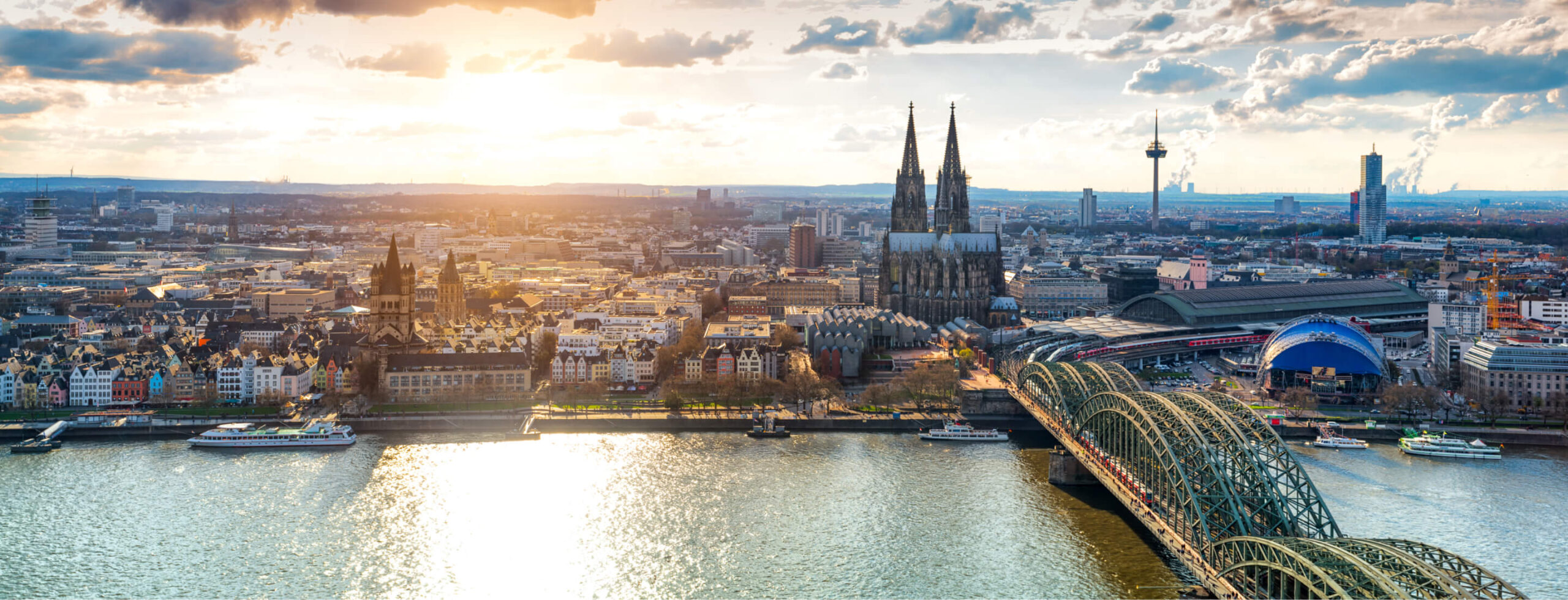Weitwinkelaufnahme der Kölner Skyline bei Sonnenuntergang. Im Zentrum des Bildes erhebt sich der Kölner Dom mit seinen beiden markanten Türmen. Die Hohenzollernbrücke überspannt den Rhein und verbindet das linke mit dem rechten Rheinufer. Mehrere Schiffe sind auf dem Fluss zu sehen. Die Stadt erstreckt sich bis zum Horizont unter einem Himmel mit vereinzelten Wolken und warmem Abendlicht.