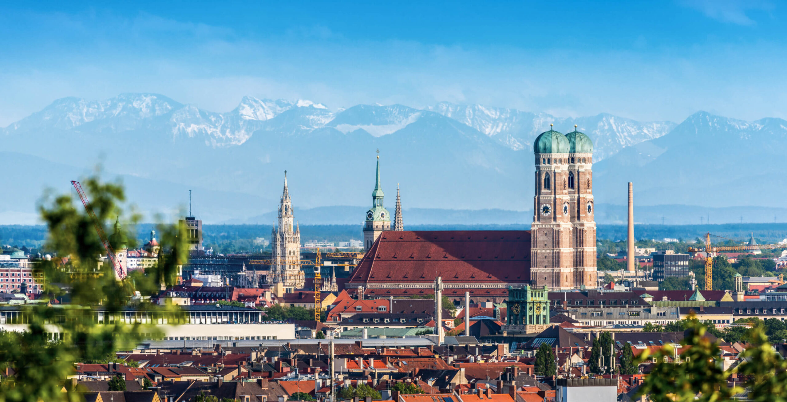 Weitwinkelaufnahme der Münchner Skyline mit der Frauenkirche im Vordergrund. Die beiden charakteristischen Türme der Frauenkirche dominieren das Stadtbild. Im Hintergrund erstreckt sich die beeindruckende Kulisse der Alpen mit schneebedeckten Gipfeln. Die Stadt München breitet sich mit ihren roten Dächern und verschiedenen Gebäuden zwischen der Kirche und den Bergen aus. Der Himmel ist blau mit einigen weißen Wolken.